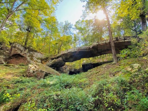 A natural stone arch surrounded by lush green trees and sunlight filtering through the foliage.