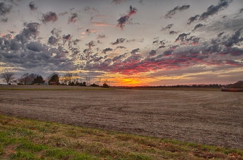 A vibrant sunset over a plowed field, with dramatic clouds and a hint of trees in the background.