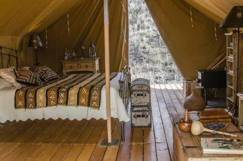 Cozy tent interior with a bed, wooden floor, decorative textiles, and rustic furnishings. Natural light filters in.