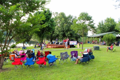 A crowd sits in colorful chairs by a lake, enjoying a performance on a stage near a pavilion surrounded by trees.