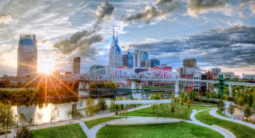 Sunset over Nashville skyline with a river, bridge, and green park in the foreground, showcasing vibrant city life.