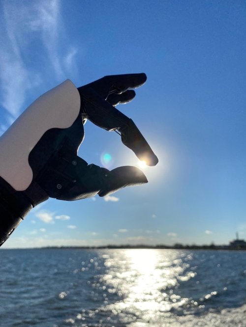 A robotic hand holds a small sun against a bright blue sky over a shimmering water surface.