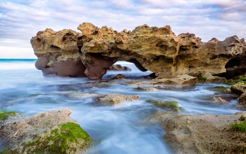 A rocky shoreline with a natural arch, surrounded by gentle waves and a cloudy sky.