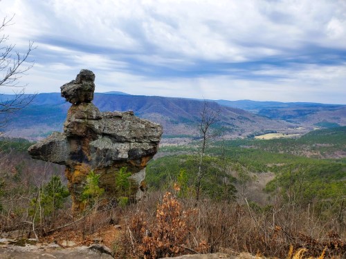 A unique rock formation stands on a hillside, overlooking a valley with rolling mountains under a cloudy sky.