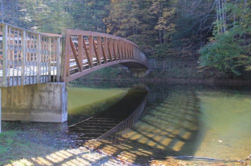 A wooden bridge arches over a calm, reflective pond surrounded by trees in autumn foliage.