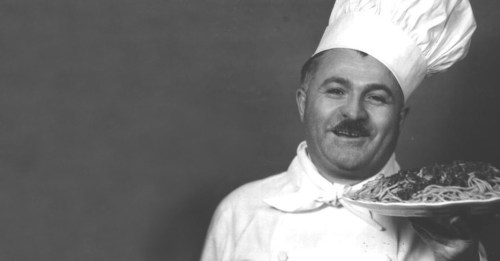 A smiling chef in a white uniform and tall hat holds a plate of spaghetti, posing against a plain background.