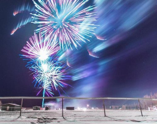 Colorful fireworks burst in the night sky over a snowy landscape, with a fence in the foreground.