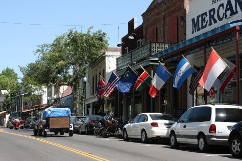 A street lined with shops and parked cars, featuring various flags hanging from buildings.