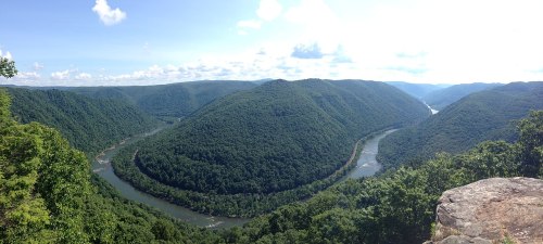 A panoramic view of lush green mountains surrounding a winding river under a bright blue sky with scattered clouds.