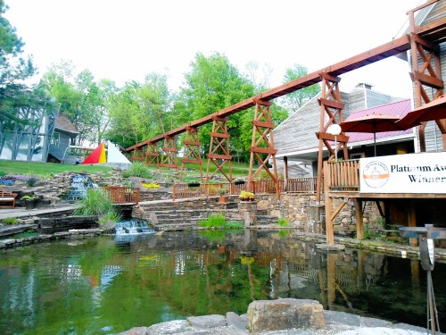 A scenic view of a wooden structure over a pond, surrounded by greenery and colorful flowers.