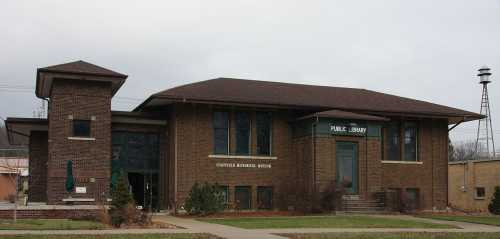 Historic brick building with a sloped roof, labeled "Public Library," surrounded by greenery and a cloudy sky.