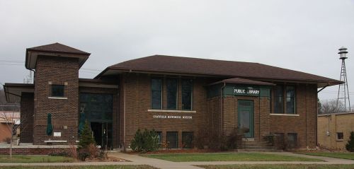 Historic brick building with a sloped roof, labeled "Public Library," surrounded by greenery and a cloudy sky.