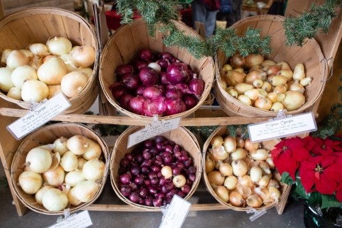 Baskets of various onions, including yellow, red, and white, displayed at a market with festive decorations.