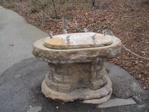 A weathered stone drinking fountain with three spouts, surrounded by a path and natural foliage.