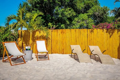 Two lounge chairs and a small table on a sandy beach, surrounded by greenery and a bamboo fence.