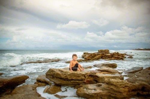 A young boy sits on rocky shore, with waves crashing and a cloudy sky in the background.