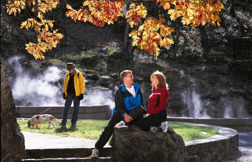 A couple sits on a rock, smiling at each other, while a person walks a dog nearby amidst autumn leaves and steam.