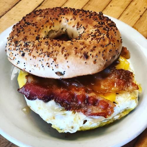 A toasted bagel topped with bacon, cheese, and a fried egg on a white plate.