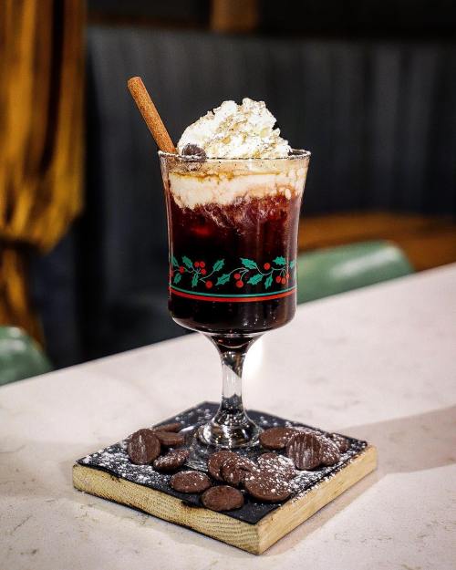 A festive drink topped with whipped cream and a cinnamon stick, served on a slate with chocolate candies.