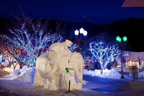 A snow sculpture of an elephant and a bear, surrounded by colorful holiday lights in a winter setting.
