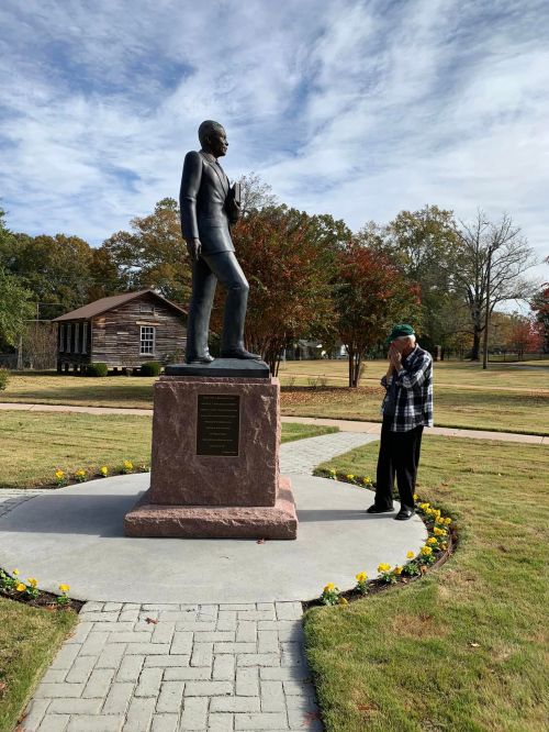 A person stands near a statue of a man in a park, surrounded by flowers and trees under a cloudy sky.