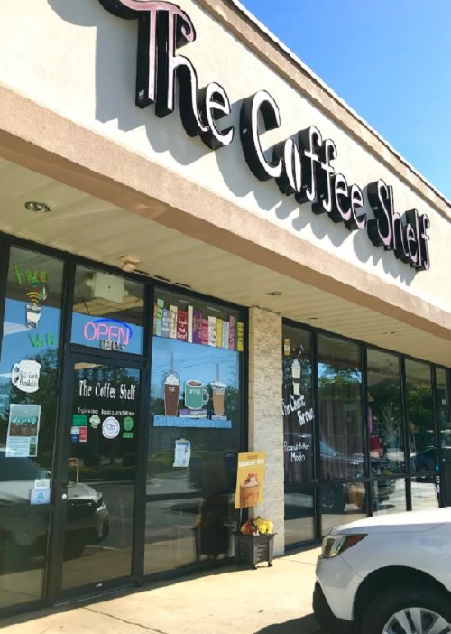 Exterior of "The Coffee Shelf" with large windows, an open sign, and colorful posters on display.