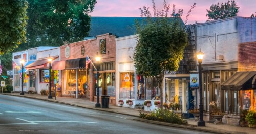 Charming small-town street lined with shops, trees, and lampposts, illuminated at dusk with a colorful sky.