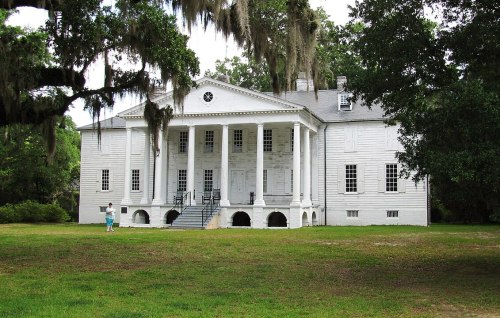 A historic white plantation house with columns, surrounded by greenery and Spanish moss. A person walks nearby.