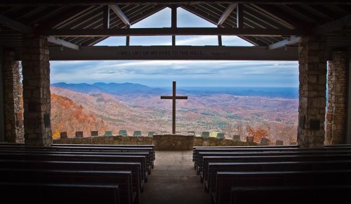 A stone chapel with a cross overlooks a scenic mountain view, framed by wooden beams and empty pews.