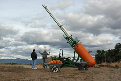 Two men stand near a large pumpkin cannon on a farm, with pumpkins in a wheelbarrow and cloudy skies in the background.