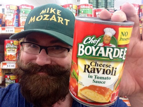 A man with a beard and glasses holds a can of Chef Boyardee Cheese Ravioli, smiling in a grocery store aisle.