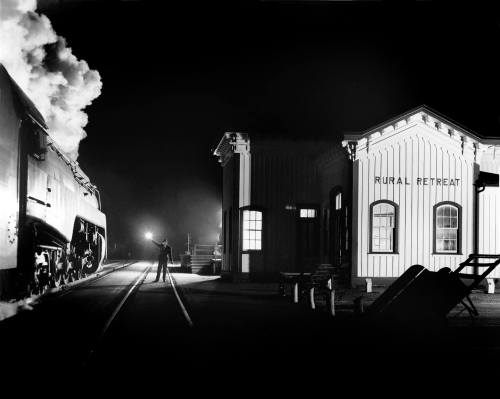 A steam locomotive at night near a station labeled "Rural Retreat," with a figure holding a lantern on the platform.