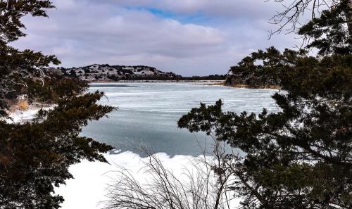 A frozen lake surrounded by trees and hills under a cloudy sky. Snow covers the ground, creating a serene winter scene.