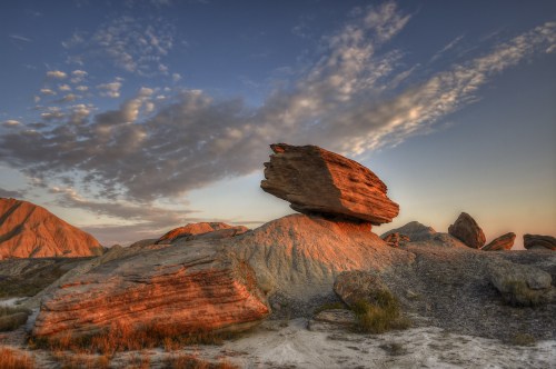 A large, balanced rock sits atop a sandy hill, illuminated by warm sunset light against a cloudy sky.