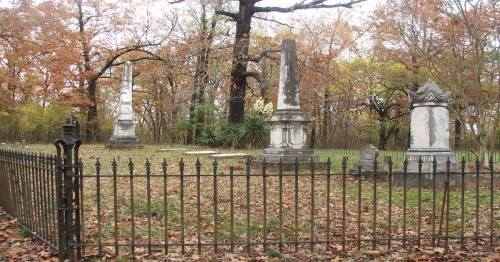 A cemetery with tall, ornate gravestones surrounded by a black iron fence and autumn foliage.