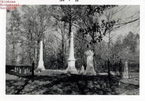 Black and white photo of a cemetery with tall monuments and a wrought iron fence, surrounded by trees.