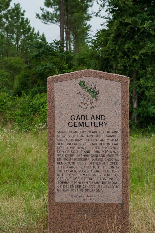 A granite marker for Garland Cemetery, detailing its history and notable graves, surrounded by trees and grass.