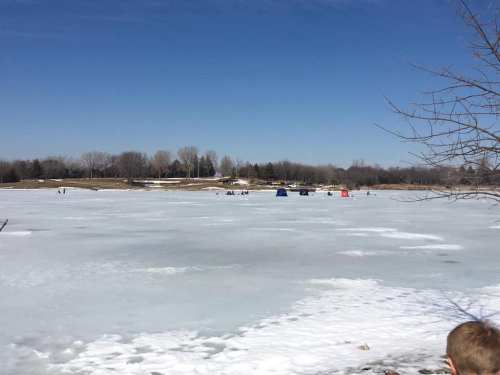 A frozen lake with patches of snow, featuring ice fishing huts in the distance under a clear blue sky.