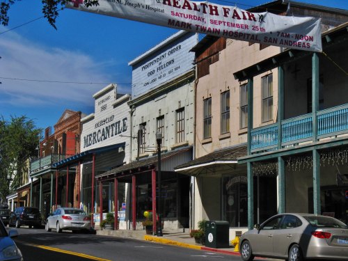 Historic storefronts line a street, featuring a mercantile and a health fair banner overhead. Cars parked along the road.