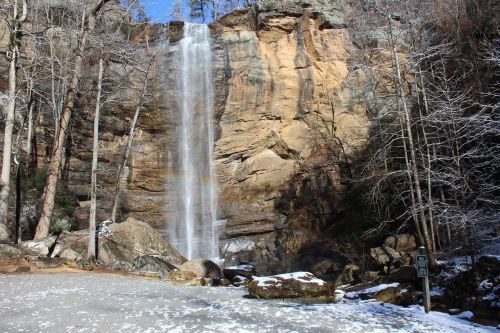 A tall waterfall cascades down a rocky cliff, surrounded by trees and a snowy ground. A rainbow is visible in the mist.