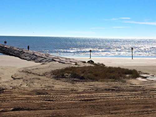 A sandy beach with a wooden pier extending into the sparkling ocean under a clear blue sky.