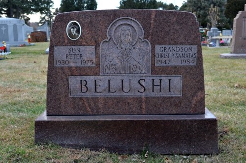 A granite gravestone with the name "Belushi," featuring engravings of a figure and details about Peter and Christ P. Samatas.