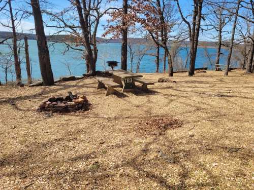A picnic table near a lake, surrounded by trees and a fire pit on a gravelly ground under a clear blue sky.
