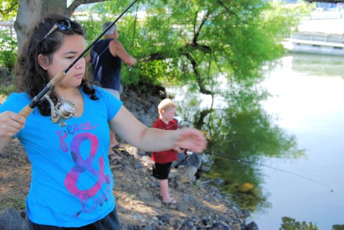 A girl in a blue shirt fishes by the water while a young boy stands nearby, both enjoying a sunny day outdoors.