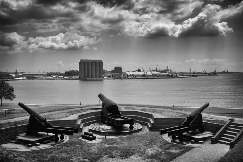 Historic cannons overlook a harbor, with dramatic clouds and industrial buildings in the background. Black and white image.