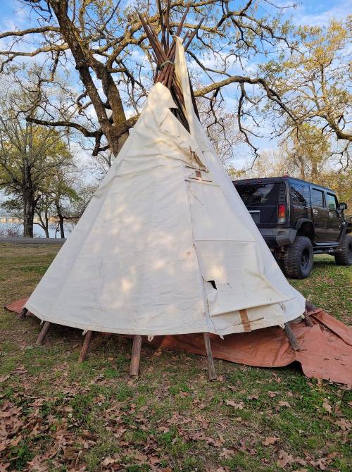 A white teepee made of fabric and wooden poles, set in a grassy area with a black SUV parked nearby.