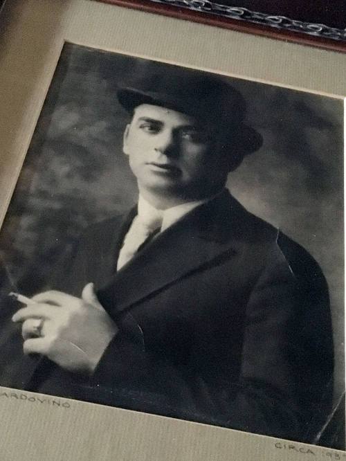 A vintage black-and-white portrait of a man in a suit and hat, holding a cigarette, circa 1920s.