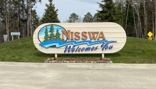 Sign welcoming visitors to Nisswa, featuring trees and water graphics, set against a green landscape.