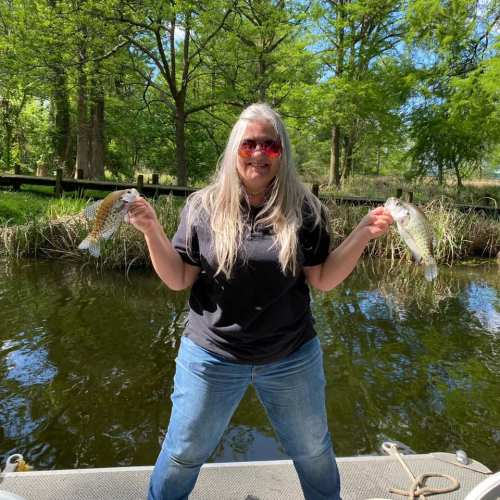 A person with long hair and sunglasses holds two fish while standing by a pond surrounded by greenery.