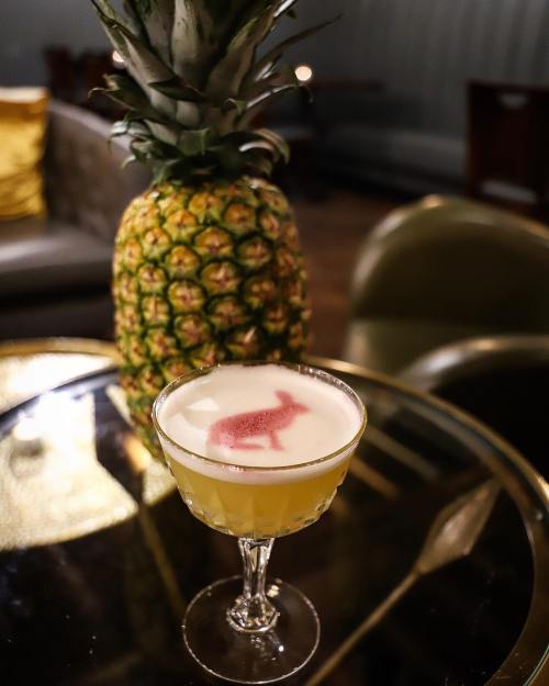 A cocktail with a frothy top and a pink design sits on a table next to a pineapple in a cozy bar setting.